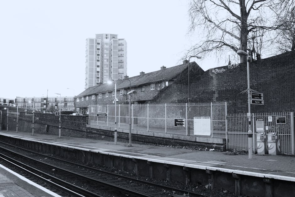 A black-and-white photograph of Barnes Cray railway station showing train tracks in the foreground, with a paved platform along the side. Behind the platform, there is a tall, multi-story residential building with numerous windows and balconies. Adjacent to the tracks, a brick wall with graffiti partially obscures a row of small, lower houses with tiled roofs. Several street lights and lampposts are positioned along the platform, and there is a metal fence with a small gate separating the station from the residential area. The overall scene captures an urban setting typical of a home relocation or furniture transport operation, with the station structure and nearby housing situated for efficient moving and unloading processes. The environment appears quiet and well-lit with outdoor lighting, suitable for loading or unloading household belongings for a move or internal house transfer.