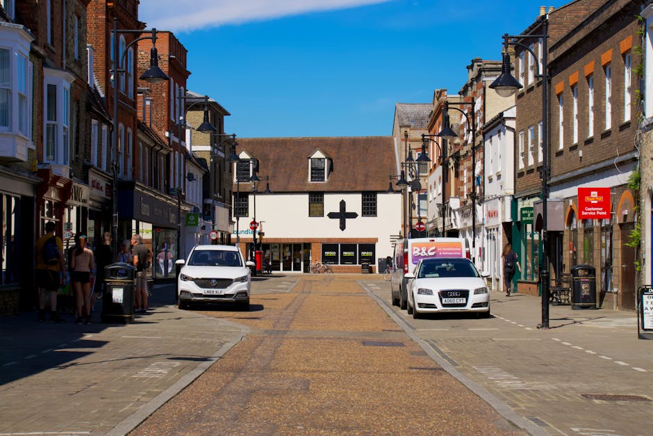 A view of a busy town centre on a bright day, featuring a mix of historic and modern buildings along Erith High Street. The scene includes pedestrians walking on the pavement, some carrying shopping b