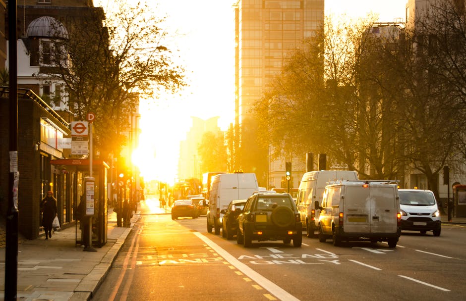 A busy urban street scene during sunset with a row of parked white commercial vans and cars in the foreground, situated next to a sidewalk with pedestrians and a bus stop shelter. The vans are positio